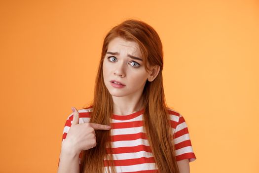 Offended innocent timid redhead girl hurt feelings, hear upsetting accusations, pointing herself frowning, look insulted, grimace frustrated, stand uneasy bothered orange background
