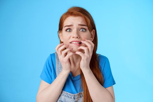 Close-up intense panicking redhead woman watching scary movie feel pressured, biting lip worried, hold hands near mouth self-soothing trying calm down, stare camera anxious, blue background
