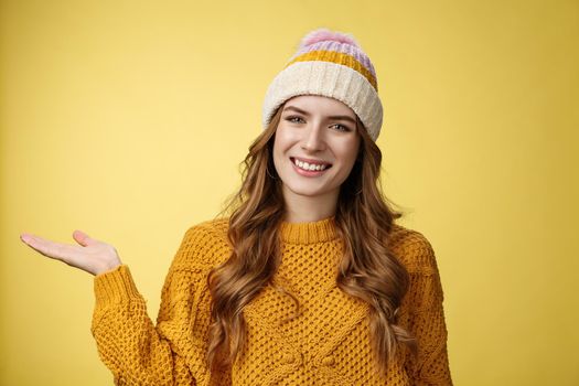 Friendly-looking confident pleasant attactive young european woman wearing winter hat, sweater extend arm show product welcoming inviting guests come in smiling happily, promoting copy space