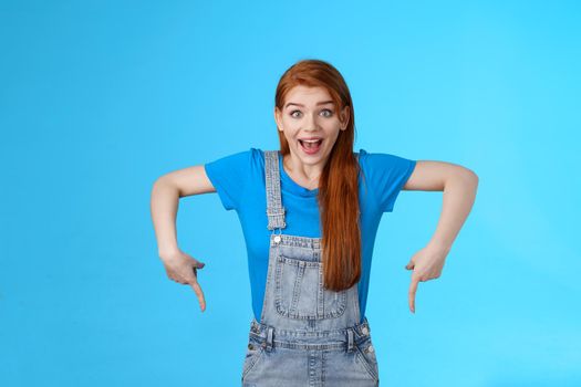 Amused wondered excited cute 20s european woman, pointing down, bottom copy space, look amazed admiration camera, describe amazing product, recommend site, stand blue background