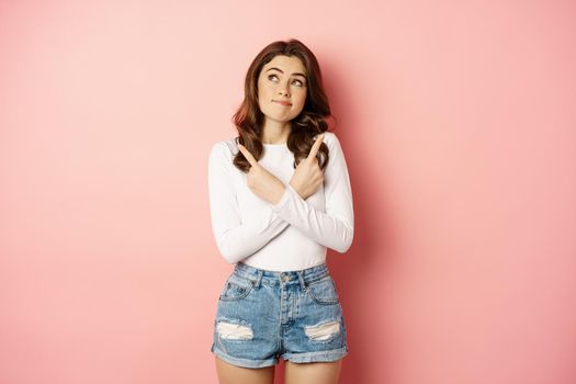 Complicated woman looking indecisive, stylish girl pointing fingers sideways and gazing thoughtful at upper left corner, making choice, deciding, pink background