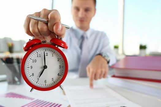 Man hand on red alarm clock stands at desk in office showing seven o'clock AM PM