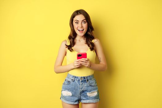 Cellular technology and online shopping concept. Enthusiastic pretty girl standing with smartphone and smiling, standing against yellow background