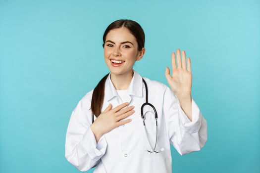 Friendly smiling woman doctor raising hand, name herself, introducing, standing in lab white coat against torquoise background