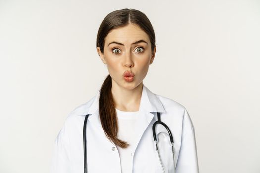 Portrait of woman doctor looking surprised, amazed, reaction of interest and amusement, standing in hospital uniform over white background