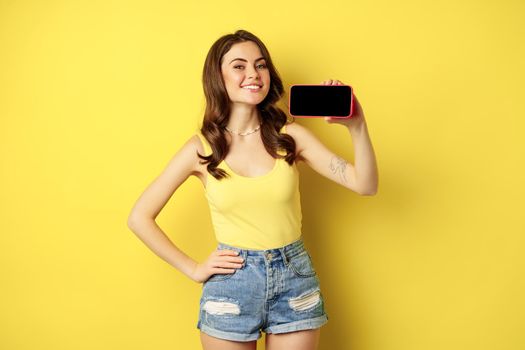 Proud smiling woman showing mobile phone horizontal screen, looking proud and satisfied, demonstrating smartphone display, app or online shopping store, yellow background