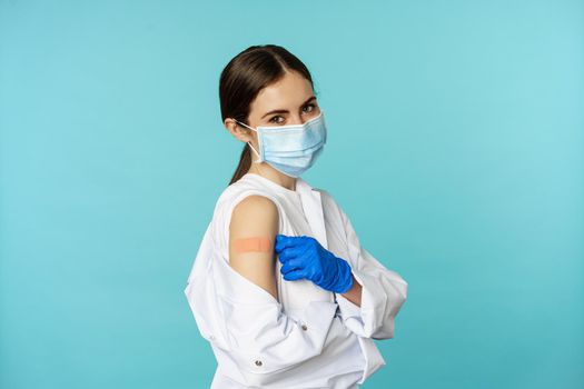 Young female doctor, nurse in medical face mask and hospital uniform, showing thumbs up after getting covid-19 omicron vaccine, showing patch on shoulder, blue background