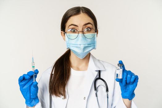 Covid-19 vaccination and healthcare concept. Young doctor in medical mask showing syringe and vaccine from coronavirus omicron variant, standing over white background