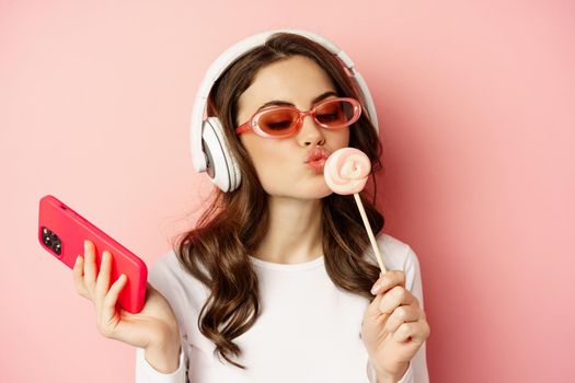 Close up portrait of stylish glamour girl, beautiful woman in headphones, listening music with smartphone, wearing sunglasses and licking lolipop