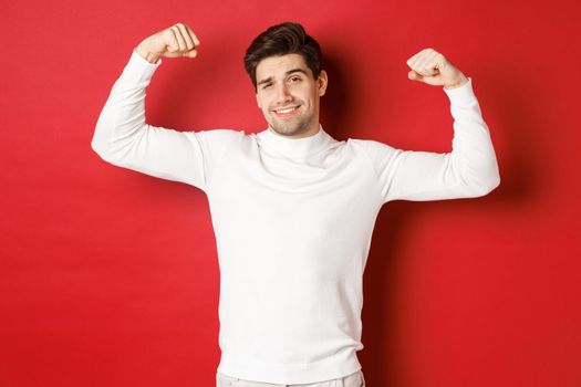 Portrait of smiling handsome man in white sweater, flexing biceps and bragging with strength, show-off strong muscles after workout, standing over red background