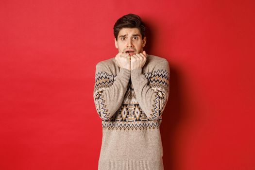 Portrait of scared and timid man in christmas sweater, looking frightened, watching something scary on new year, standing over red background