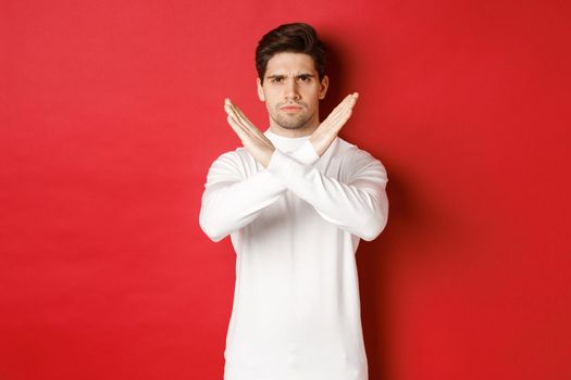 Image of serious and angry man in white sweater, express storng disapproval, showing cross sign to stop something bad, forbid action, standing over red background
