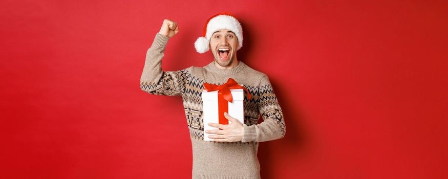 Image of happy and excited handsome man, reicing christmas gift, raising hands up in triumph and smiling, celebrating new year, standing over red background in santa hat