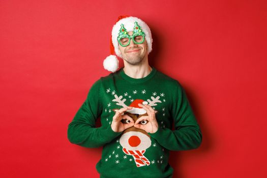 Image of happy smiling man in party glasses and santa hat, fooling around with funny christmas sweater, celebrating winter holidays, standing over red background