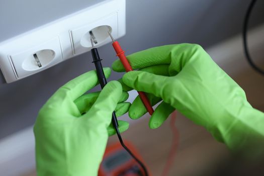 Hands in green gloves plug voltmeter into an outlet in wall
