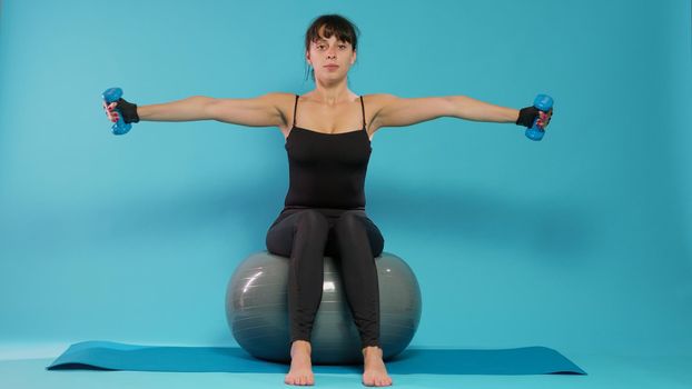 Athlete lifting dumbbells and sitting on fitness toning ball in studio, training arms muslces with sport weights and equipment. Active woman exercising and stretching with workout activity.