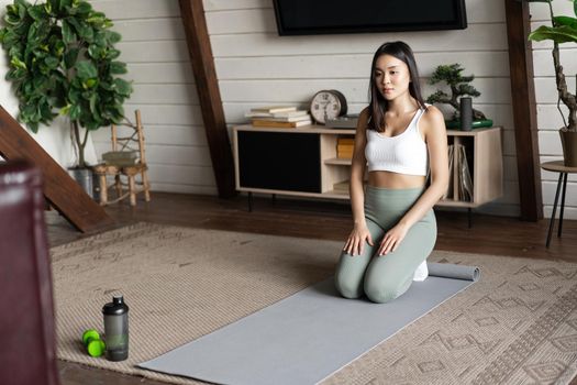 Image of young asian woman doing yoga at home on floor mat, meditating in activewear in living room