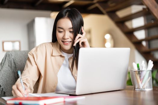 Smiling asian girl working from home, talking with client, answer phone call and taking notes in notebook, sitting near laptop