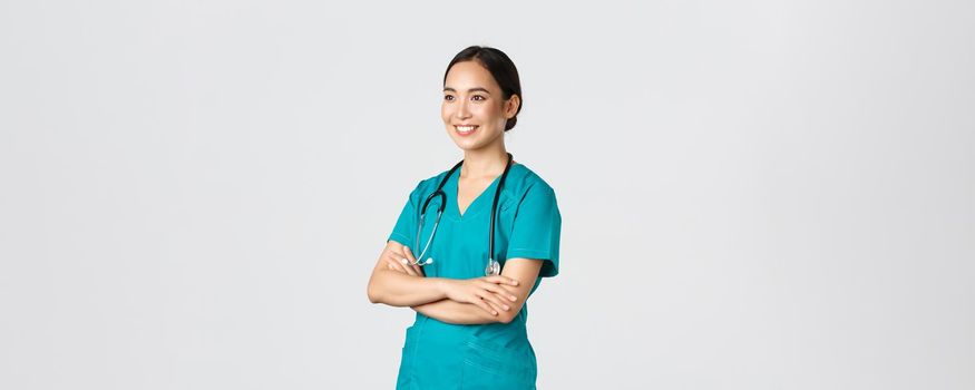 Covid-19, healthcare workers, pandemic concept. Side view of professional confident and hopeful asian female doctor, nurse looking assured away and smiling, standing in scrubs white background