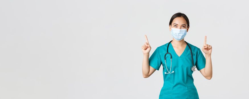 Covid-19, coronavirus disease, healthcare workers concept. Pleased cunning asian female nurse, doctor in medical mask and scrubs, looking and pointing fingers up, white background