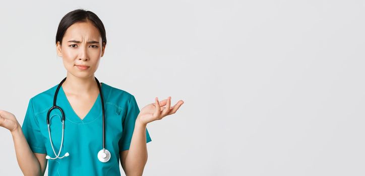 Healthcare workers, preventing virus, quarantine campaign concept. Close-up of confused and puzzled asian female physician in scrubs having complicated problem, raising hands and shrugging