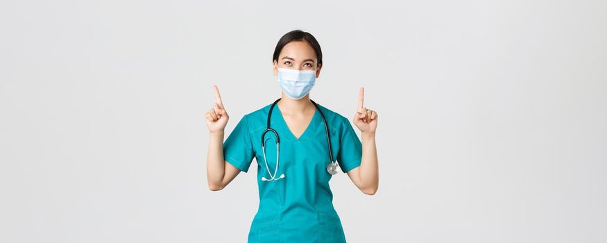 Covid-19, coronavirus disease, healthcare workers concept. Pleased cunning asian female nurse, doctor in medical mask and scrubs, looking and pointing fingers up, white background
