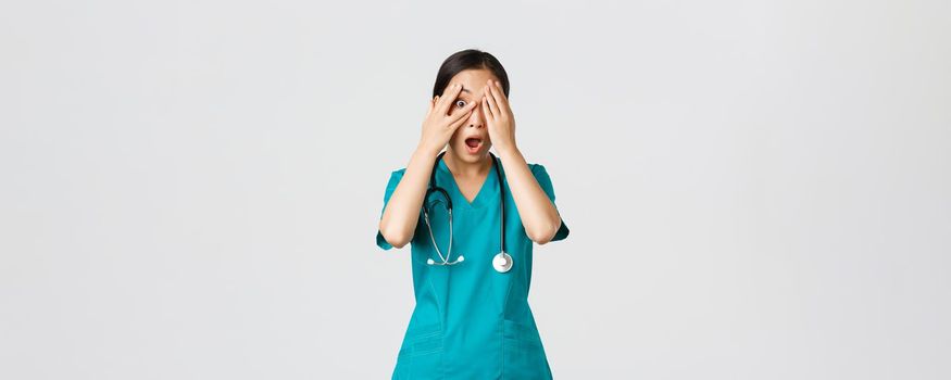 Covid-19, healthcare workers and preventing virus concept. Scared and shocked asian female nurse, doctor in scrubs witness something embarrassing or scary, cover eyes but peek through fingers