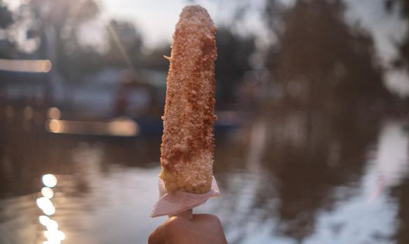 Hand holding traditional corn with mayonnaise and chili powder above lake