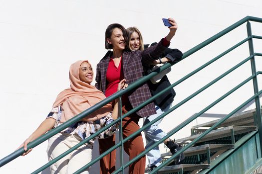 Cheerful multiracial girlfriends taking selfie on smartphone on stairs