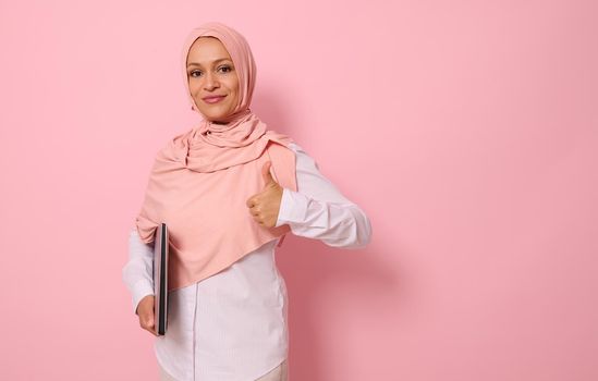 Confident portrait of a successful gorgeous Arab Muslim woman in pink hijab and strict casual attire showing a thumb up, looking at camera, posing against colored pastel background with space for text