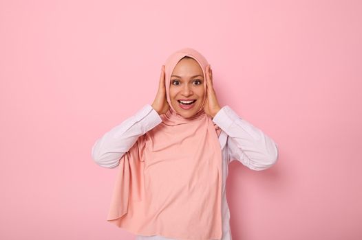 Happy religious Muslim woman in pink hijab holding her head with hands and smiling with toothy smile looking at camera with surprised look, isolated on colored background with copy space.