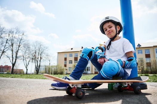 Bottom view of smiling boy in protective gear of skateboarder sitting on skateboard and looking at camera