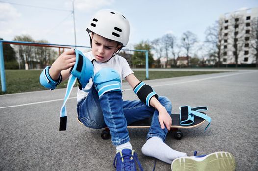 Adorable boy skateboarder in safety helmet and gear sitting on wooden skateboard on playground asphalt