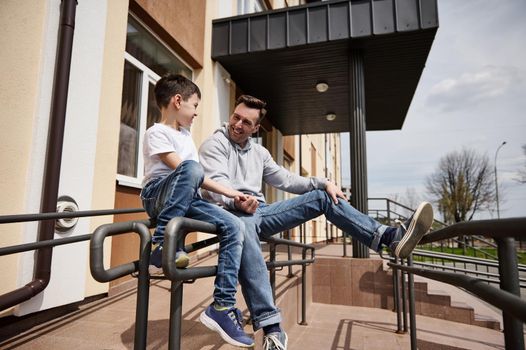 Happy young father sitting on the handrail and happily communicating with his son on a warm spring day