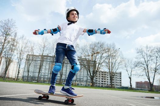 Happy and smiling boy in protective gear and helmet keeps balance while riding a skateboard