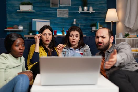 Portrait of multi-ethnic friends sitting on couch watching online movie on laptop computer enjoying hanging out together. Group of people enjoying relaxing time in living room. Friendship concept