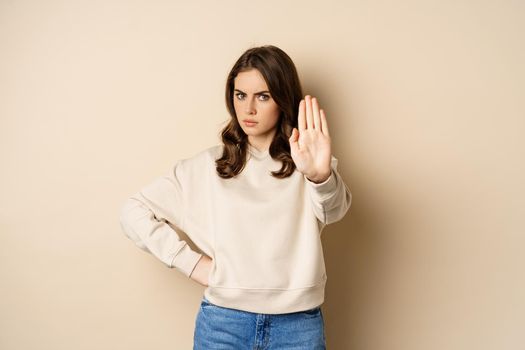 Stop. Serious and confident woman showing extended arm palm, prohibit, forbid smth, blocking something, standing over beige background