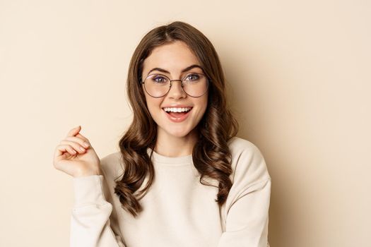 Close up portrait of stylish brunette woman in glasses, laughing and smiling, posing in eyewear against beige background