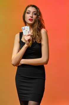 Brunette female with earring in nose, in black dress. Showing two playing cards while posing on colorful background. Poker, casino. Close-up