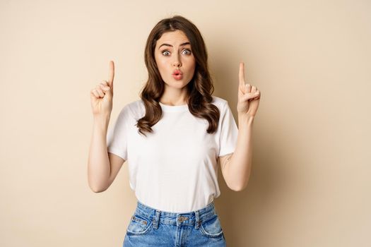 Excited girl looking interested, pointing fingers up, showing advertisement, standing in tshirt over beige background