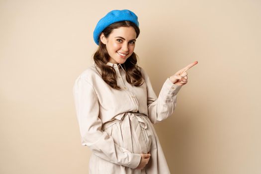 Young future mother, pregnant woman smiling, posing with big belly third trimester of pregnancy, looking happy, expecting baby, pointing right at logo, banner or advertisement