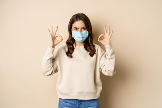 Covid-19, pandemic and quarantine concept. Young woman wears medical face mask during coronavirus omicron outbreak, showing okay sign, standing over beige background