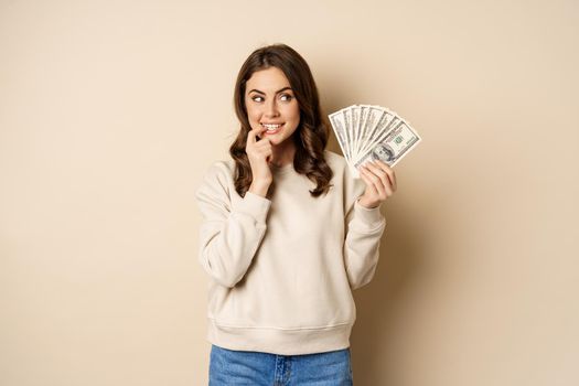 Excited brunette woman holding money and biting finger with yearning face, watns to buy smth, shopoholic with cash, standing over beige background