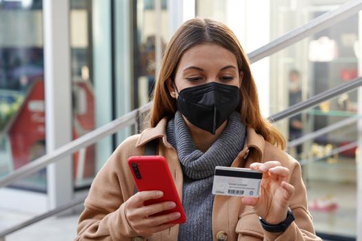 Credit card payment outdoors. Beautiful young woman with black protective mask enters her credit card number on smart phone for online shopping sitting in city.