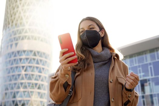 Portrait of young business woman wearing protective mask FFP2 KN95 walking in modern city street using her telephone