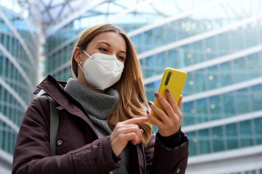 Portrait of woman wearing protective medical face mask using a phone with modern city background. Low angle.