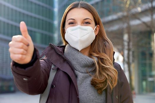 Close up of optimistic business woman wearing protective mask KN95 FFP2 showing thumbs up in modern city street and looking at camera