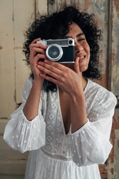 Portrait of young multiracial woman holding vintage style camera taking a picture looking at camera. Vertical image.