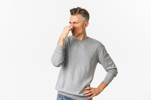 Portrait of handsome middle-aged man shut nose from disgusting smell, complaining on stink, standing over white background