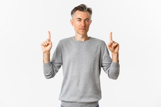 Image of thoughtful and unsure adult man, squinting hesitant while pointing fingers up, making choice, standing over white background